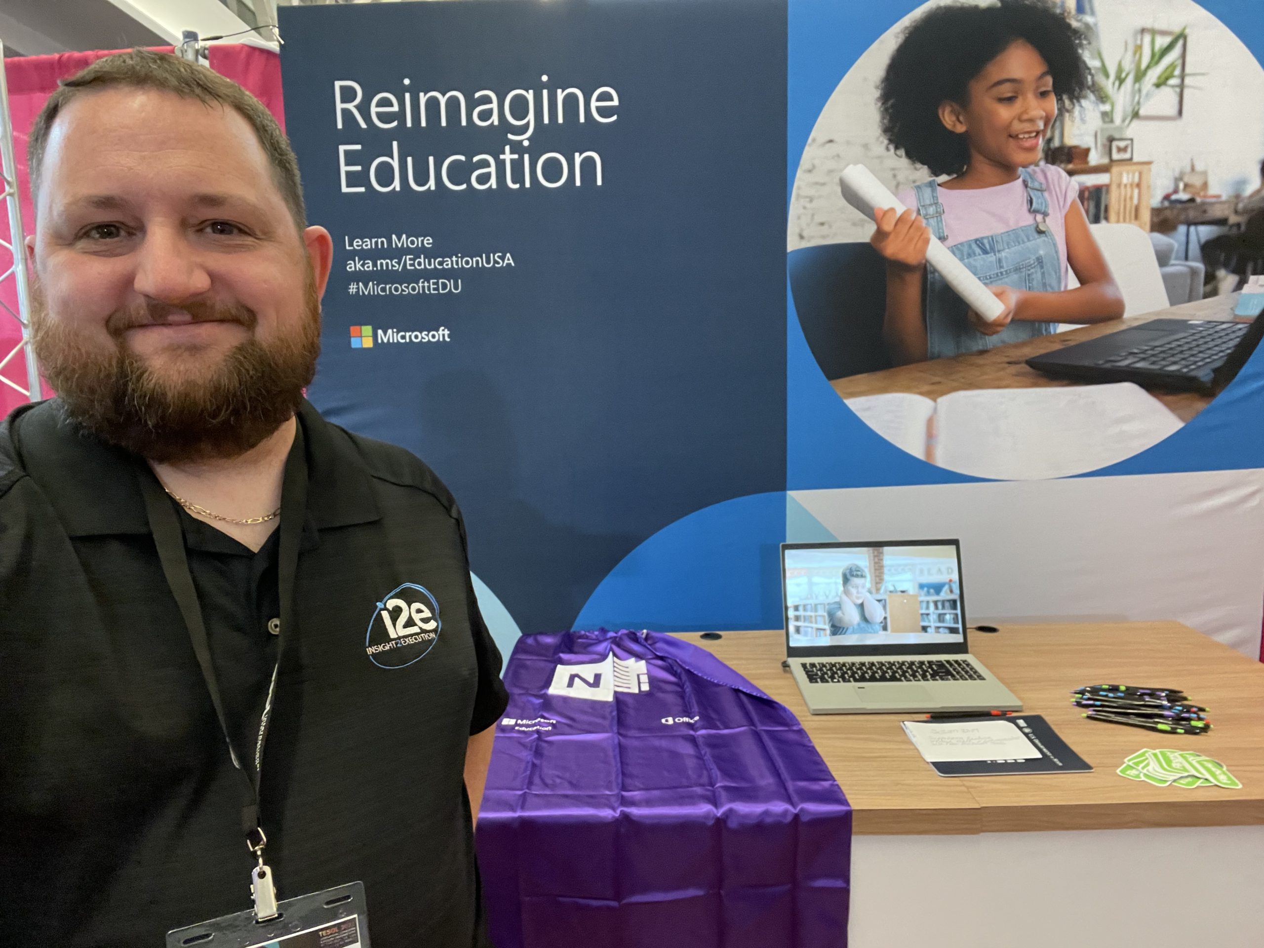 Seth Fewell, working the Microsoft booth at the TESOL Conference on behalf of i2eEDU, wearing his i2eEDU logoed shirt, with a OneNote cape, a Surface Laptop Studio, pens, & stickers on a table in the background.